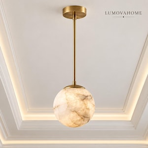 May include: A pendant light fixture featuring a spherical shade made of translucent, marbled stone with beige and cream veining. The shade is suspended by a thin, vertical brass rod connected to a circular brass ceiling canopy. Text reads LUMOVAHOME.