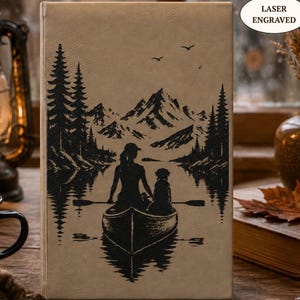 Può includere: Un taccuino rettangolare color marrone chiaro con un'illustrazione nera incisa al laser di una persona e un cane in canoa su un lago, circondati da pini e montagne. Il testo LASER ENGRAVED è stampato in un ovale bianco in alto a destra.