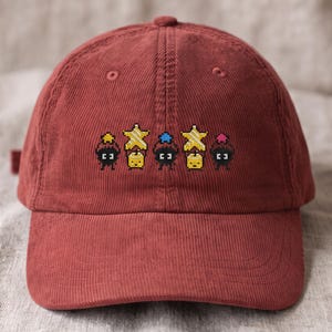 May include: A dark red corduroy baseball cap featuring an embroidered pixel art design. The embroidery displays five small black soot-like figures, two yellow star shapes, and three small colored hats in yellow, blue, and pink, arranged in a row.