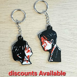 Op de afbeelding: Twee zwart-witte acryl sleutelhangers met zijprofielen van twee personen met rode spetterpatronen op hun gezicht. Elke sleutelhanger bevat een zilveren metalen ring en ketting. De tekst "discounts Available" is zichtbaar.