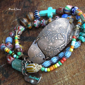 Może przedstawiać: Bransoletka wielorzędowa z zawieszką z miedzi w kształcie drzewa. Bransoletka wykonana jest z różnych kolorowych szklanych koralików, w tym turkusowych, niebieskich, czerwonych, pomarańczowych, żółtych i zielonych. Zawieszka jest grawerowana tekstem "Rark&Jew".