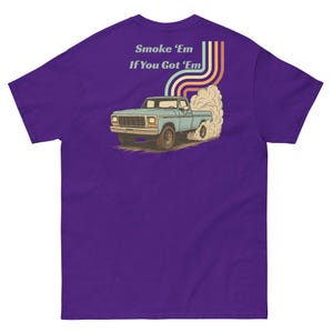 May include: A purple t-shirt featuring a graphic on the back of a light blue pickup truck performing a burnout with a large cloud of smoke. Above the truck, colorful rainbow-colored lines curve upward with the text Smoke 'Em If You Got 'Em.