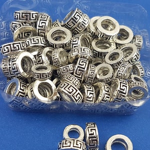 May include: A close-up of a clear plastic container filled with silver metal beads. The beads are round and have a Greek key design. There are five beads outside of the container.