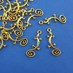 Può includere: Un gruppo di ciondoli a forma di lucertola in metallo dorato con la coda arricciata. I ciondoli sono sparsi su uno sfondo blu.