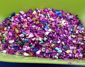 以下が含まれることがあります： 虹色で金属光沢のあるカラフルなガラスの破片の山をクローズアップした写真。破片は主にピンクで、ゴールド、ブルー、グリーンのアクセントが加えられています。