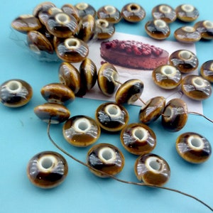 May include: A collection of round, doughnut-shaped ceramic beads with a glossy brown and cream glaze. Each bead features a central hole and a smooth, reflective surface. Some beads are strung on a thin wire, while others are scattered loosely.