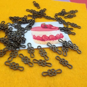 May include: Bronze-colored metal filigree connectors scattered on a yellow surface. The connectors have a decorative, openwork design with multiple loops and circles. A picture of a strawberry cake is in the background.