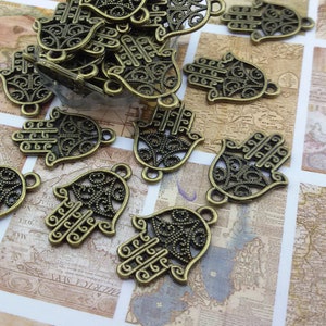 May include: A pile of antique gold-toned metal charms in the shape of the Hamsa hand, a symbol of protection and good luck. The charms are intricately detailed with filigree designs.