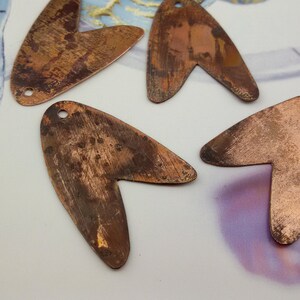 May include: Four copper-colored metal pendants in the shape of a heart with a pointed tip. The pendants have a rustic, aged look and are ready to be used in jewelry making.