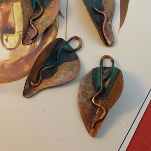 Puede incluir: Cuatro colgantes de cobre en forma de hoja con una pátina verde y un detalle de alambre retorcido. Los colgantes tienen un agujero en la parte superior para colgar.