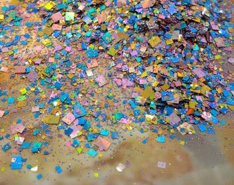 May include: A pile of iridescent square glitter in shades of gold, blue, pink, and purple. The glitter is scattered on a light brown surface.