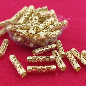 以下が含まれることがあります： 金色の円筒形ビーズのコレクション。複雑な透かし彫りのデザインが施されています。ビーズは赤い表面に散らばっており、透明なビニール袋からこぼれ落ちているものもあります。これらの装飾ビーズは、ジュエリー作りや工芸に使用される可能性があります。