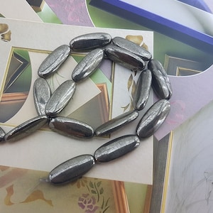 May include: A strand of oval-shaped, dark gray pyrite beads. The beads have a metallic sheen and are arranged in a loose circle.