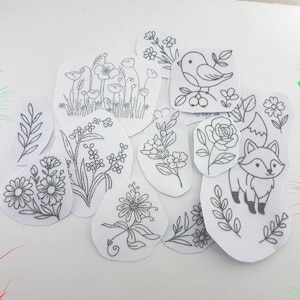 Peut inclure: Un ensemble de patchs en tissu blanc avec des motifs brodés. Les patchs présentent divers motifs floraux et animaliers, dont un oiseau, un renard et plusieurs compositions florales. La broderie est réalisée avec un fil gris foncé, créant un effet de contraste.