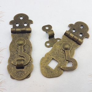 May include: Two antique bronze-coloured latches with ornate floral designs. Each latch has a hinged top with three holes, a decorative body, and a hook-and-eye closure. Suitable for vintage boxes or decorative projects.