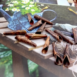 May include: A collection of 14 copper-coloured metal tubes with a spiral design. The tubes are arranged on a wooden surface with a blue and white watering can and green foliage in the background.