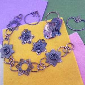 Könnte beinhalten: Ein Set aus Kupferschmuck, bestehend aus einer Halskette, Ohrringen und einem Ring. Die Stücke sind alle im Blumendesign gehalten und haben einen rustikalen, Vintage-Look. Der Schmuck wird auf einem gelben, grünen und violetten Filz-Hintergrund präsentiert.