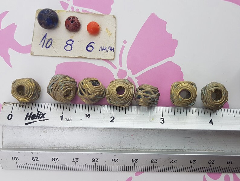 May include: Eight gold wire wrapped beads, each approximately 1 inch in diameter, are lined up on a ruler. The ruler is marked in inches and 1/16ths of an inch. A small card with the numbers 10, 8, and 6 is visible in the background.