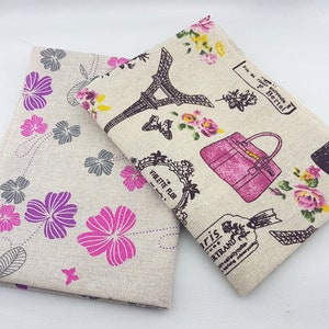 May include: Two beige linen fabric squares with different patterns. One square has a pink floral pattern with a pink handbag and Eiffel Tower. The other square has a purple and pink floral pattern.