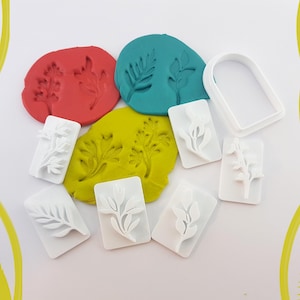 Puede incluir: Vista cenital de cortadores de galletas blancos con forma de hoja y plastilina de colores. La plastilina es roja, turquesa y amarilla, con impresiones de hojas. Los cortadores son rectangulares y presentan varios diseños de hojas.