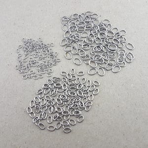 Könnte beinhalten: Eine Ansammlung kleiner, silberfarbener, ovaler Biegeringe, die auf einer grauen Oberfläche verstreut sind. Die Biegeringe sind in drei separaten Haufen angeordnet, die in Größe und Menge variieren. Diese Schmuckherstellungskomponenten sind einsatzbereit.