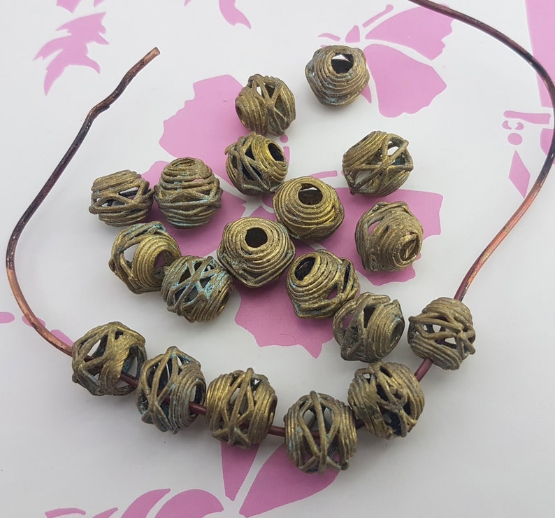 May include: A collection of 18 gold-toned metal beads with a wire mesh design. The beads are arranged in a circle on a pink background.