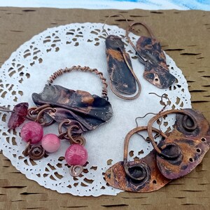 May include: A selection of handcrafted copper earrings and a pendant with pink beads. The earrings display distinctive shapes and textures, with a blend of dark and light copper hues. The jewellery is presented on a white doily.