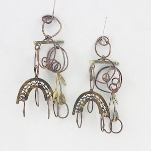 May include: A pair of gold and copper earrings with a geometric design. The earrings feature a curved, openwork piece with a delicate pattern, a small, flat, rectangular piece, and several loops and swirls of wire.