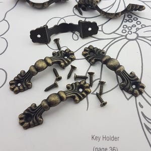 May include: Antique bronze-colored cabinet pulls and screws are scattered on a floral patterned surface. The pulls have an ornate, decorative design. The image also includes a small, curved handle and the text "Key Holder (page 36)."