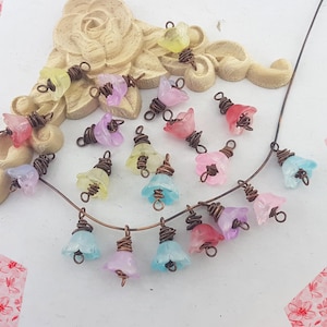 May include: A selection of translucent flower-shaped beads in various colours, including pink, purple, yellow, and blue, with copper wire detailing. Some are strung on a thin copper wire, others scattered near a decorative beige element.