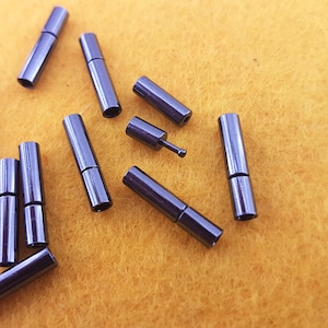 Könnte beinhalten: Nahaufnahme von zehn schwarzen Metall-Verbindungsstücken auf einer gelben Oberfläche. Die Verbindungsstücke sind zylindrisch und haben an einem Ende einen kleinen Metallstift.