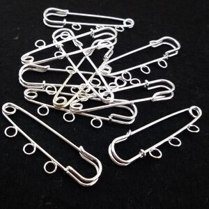 Puede incluir: Un grupo de imperdibles plateados con múltiples bucles en cada imperdible. Los imperdibles están dispuestos en una pila sobre una superficie negra.