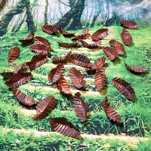 Könnte beinhalten: Eine Sammlung von braunen, metallischen, blattförmigen Cupcake-Förmchen. Die Förmchen sind auf einem grün-braunen Hintergrund verstreut, der einem Waldweg ähnelt.
