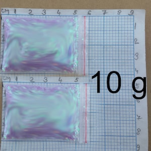 以下が含まれることがあります： 虹色のグリッターが詰まった透明な小さなプラスチック袋2つ。各袋には10グラムのグリッターが入っています。袋はグリッド状の表面に置かれています。