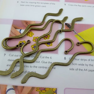 May include: Five antique gold-toned metal hooks with a wavy design. The hooks are arranged in a pile on a pink and white surface.