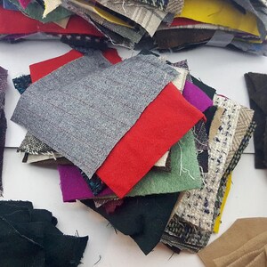 Op de afbeelding: Een verzameling stofstalen in verschillende kleuren en texturen. De stalen bevatten grijze, rode, groene, paarse en bedrukte stoffen. De stoffen zijn in een stapel gerangschikt, waarbij de verschillende materialen worden getoond.