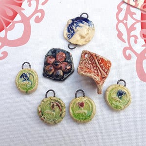 Può includere: Una collezione di ciondoli in ceramica fatti a mano in varie forme e design. I ciondoli presentano motivi floreali e astratti, con colori tra cui blu, rosso, verde e marrone. Ogni ciondolo ha un piccolo anello di metallo per appenderlo.