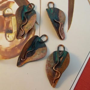 Op de afbeelding: Vier blad-vormige hangers met een koperen afwerking, geaccentueerd met turquoise en bruine tinten. Elke hanger heeft een lus aan de bovenkant om op te hangen en een golvend, koperkleurig accent. De hangers zijn gerangschikt op een lichtgekleurd oppervlak.