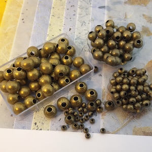 May include: A close-up of a collection of antique gold metal beads in various sizes. The beads are arranged in small containers and scattered on a textured surface.