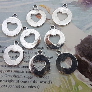 Op de afbeelding: Zeven zilverkleurige metalen bedels met een hartvormige uitsparing. Elke bedel heeft een lus aan de bovenkant om te bevestigen aan een ketting, armband of andere sieraden.
