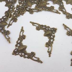 May include: Ornate, antique brass-toned metal corner brackets. Each bracket features a central teardrop shape and decorative scrollwork. These decorative elements are ideal for adding a vintage touch to craft projects, scrapbooking, or furniture restoration.