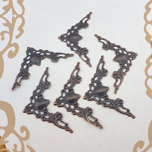 Könnte beinhalten: Eine Sammlung verzierter, bronzefarbener Metallecken. Jede Ecke weist ein dekoratives, durchbrochenes Design mit einer zentralen, soliden Schildform auf. Die Ecken sind auf einer weißen Oberfläche mit einem dezenten, gemusterten Rand angeordnet.