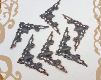 Può includere: Una collezione di angoli decorativi in metallo color bronzo. Ogni angolo presenta un design traforato ornato con una forma a scudo centrale solida. Gli angoli sono disposti su una superficie bianca con un bordo sottile.