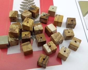 May include: A collection of 18 brown, tan, and white jasper stone beads. Each bead is a cube with a hole drilled through the center. The beads are scattered on a red and white surface.