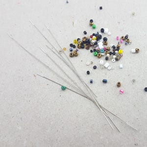Könnte beinhalten: Eine Ansammlung kleiner, farbiger Perlen und mehrere lange, dünne Nadeln, die auf einer hellgrauen Oberfläche verstreut sind. Die Perlen sind in verschiedenen Farben gehalten, darunter Gold, Schwarz, Weiß, Rosa und Grün. Die Nadeln sind silberfarben.
