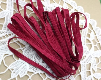 May include: A spool of burgundy red elastic with a shimmery finish. The elastic is folded and arranged on a white lace doily.