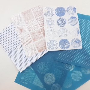 Puede incluir: Una colección de plantillas estampadas en varios diseños y colores. Las plantillas incluyen formas geométricas, puntos y patrones circulares en tonos de azul, rosa y blanco. Las plantillas están dispuestas sobre una superficie blanca.