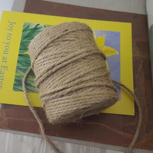 Puede incluir: Una bola de hilo natural, de color marrón claro, descansa sobre una tarjeta amarilla. La tarjeta presenta el texto "Joy to you at Easter" y un diseño floral. El hilo probablemente se utiliza para manualidades o envolver regalos.