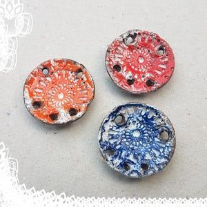 Puede incluir: Tres adornos redondos de cerámica con fondo blanco y un diseño en relieve en el centro. Los adornos están pintados en rojo, naranja y azul.