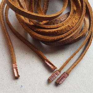 Puede incluir: Cordones de cuero marr&oacute;n enrollados con terminales de color cobre. Los cordones tienen un tono c&aacute;lido y terroso, y los terminales son rectangulares. El cuero parece texturizado y desgastado, lo que sugiere una est&eacute;tica vintage o r&uacute;stica.